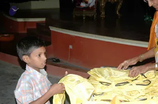 A young boy in a plaid short-sleeved shirt stands at left, reaching toward a pile of folded yellow fabric items with black trim on a table. At right, an older woman in an orange sari and gold bangles, with a wristwatch and glasses, handles the yellow garments. A raised stage with a red-and-cream base and an ornate gold-and-wood table or throne is visible in the background, with a small brass dish at left edge. The setting appears to be an indoor hall.