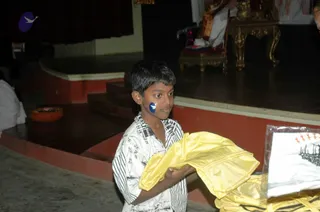 A young boy with short dark hair stands on a dark stage floor holding a folded yellow fabric garment over his arm. He wears a white shirt with black striped and printed patterns and has a small blue and white bird emblem painted on his cheek. Behind him, a raised dais with a red edge and a golden ornate seat or throne holds a figure in white draped clothing near a small brass lamp on a stand, with a dark curtain at left. Another yellow fabric bundle and a white folded cloth lie at the lower right edge of the frame.
