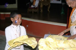 A young boy in white ceremonial kurta with a rudraksha-style mala and a blue dot on his cheek stands on the left, holding folded yellow silk garments. On the right, an older woman with glasses, a flowing white and yellow sari and an orange shawl, with beaded necklaces and a bangle, reaches toward a pile of yellow cloth and a plastic-wrapped package. Behind them a dark stage has a golden ornate chair and a small lit green tree with tinsel, and a portrait is visible at the top. The setting appears to be an ashram hall.