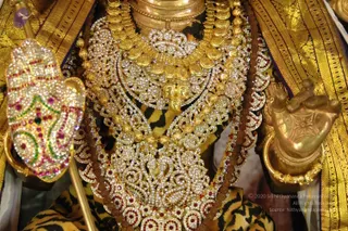 Close-up photograph of a deity murti adorned with heavy gold jewellery set with white stones and red and green gems. A layered gold necklace with ornate paisley and floral openwork covers the chest, with rudraksha beads visible beneath. A gold-and-stone hand with raised palm and a gold foot appear at the sides, with a gold-bordered zari silk cloth of yellow and black tiger-stripe pattern beneath. No people are visible.
