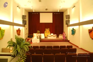 Interior of an auditorium-style hall with rows of maroon upholstered seats facing a raised stage. A large blank projection screen hangs against deep red drapes. On the stage, a golden throne-like chair with a framed portrait of a seated figure in saffron sits behind a white draped table and decorated altar with flowers. A potted fern stands in the foreground, and a woman in a pink sari walks across the stage while another person in dark clothing is partially visible at the left edge. Colourful draped sari-style wall decorations in green, orange and teal line the side walls, and boxes are stacked at the lower left.