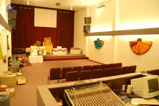 An interior view of a hall being set up as a temple or satsang space. Red curtains hang behind a raised stage with a projection screen. On the stage stands a framed portrait of a seated deity murti on a yellow cloth-covered seat, flanked by white-draped tables with flower garlands and a potted plant. Rows of maroon chairs face the stage. In the foreground a mixing console and an old computer monitor sit on a counter, and cardboard boxes lie on the floor at left. Two adults and a child sit near a stroller at the left edge. Two draped fabric pieces, one teal and one orange, hang on the white wall at right.
