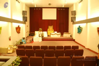 Interior of a plain auditorium-style hall with white walls, warm ceiling lights and rows of brown upholstered theatre seats facing a raised stage. Dark red curtains hang behind a blank projection screen. On the stage, a gilded framed picture sits on a tall yellow-draped chair beside a white-draped table decorated with greenery and garlands, and smaller framed pictures rest on another white-draped table. Potted plants and a bouquet stand nearby. Colourful draped fabric pieces are mounted on the side walls. At the left, a seated woman in a blue outfit tends a small child beside a stroller, and cardboard boxes sit on the floor. A few partially visible figures appear at the edges of the frame.