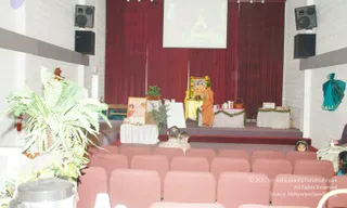 Photograph of a hall with maroon curtains and a projection screen above a stage. On the stage, a figure in orange robes stands beside a draped table with white cloth and green garland, in front of a framed portrait decorated with marigold garlands. A small table with a potted plant and a framed photo stands on the left of the stage, and a teal cloth hangs on the right wall. Rows of maroon chairs fill the foreground, with a few seated people in the audience, including a man and a child on the left and a girl on the right. Potted ferns and a metal urn sit near the front.