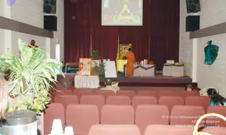 Photo of a small auditorium with rows of maroon upholstered seats, a raised stage backed by dark red curtains, and a projection screen above showing a golden temple-like structure with figures. On stage, a figure in orange robes stands beside a golden chair holding a framed portrait, with a table draped in pink and garlanded with green foliage, small containers, and a basket. A poster on an easel and a whiteboard sit to the left, with potted ferns in the foreground. Several seated people are visible in the audience, including a child in front and a person wearing headphones at the right. A green draped cloth hangs on the side wall.