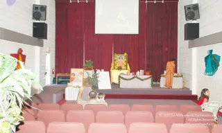 A photograph of a small hall with red upholstered seats in rows facing a raised stage draped in deep red curtains. On the stage, a framed portrait of a saffron-robed figure with a garland of flowers sits on a gold-draped table, beside a vase of greenery, a basket and small items on a white-covered table. A figure in an orange robe stands at the right side of the stage. A projection screen above shows an image. A seated adult and a child face the stage from the front row, and a young girl in red sits at the right edge. A poster stands on an easel at the left of the stage, and a blue-green wrapped cloth hangs on the right wall.