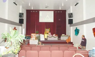 Interior of a white-walled hall with rows of pink upholstered seats facing a stage draped in deep maroon curtains. A projection screen above the stage shows a seated figure on a throne. On the stage, a person in orange robes sits on a gold-and-yellow throne-style chair beside a draped table decorated with green garland, with flowers, a small plant and a basket. A standing man in a dark sweater looks toward the stage on the right, and a person lies prostrate on the floor near the front seats. Potted ferns, a teal cloth hanging on the wall and a framed certificate on an easel complete the scene.