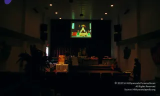 A dim auditorium photographed from the back rows looking toward a stage. A projection screen above the stage shows a gold-and-red ornate throne or altar graphic with green cushions. On stage a draped table holds a small arrangement and a lamp, with a potted plant in the foreground. Two people are visible: one seated on the right side of the stage in a red garment, and one standing at the far right edge in a dark jacket. Dark curtains and wall-mounted speakers frame the room.