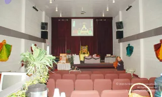 Interior of a small hall with white brick walls and a red-brown curtained stage. A projector screen above the stage shows a golden-robed seated figure on a throne. On the stage, an empty yellow-draped throne with a framed portrait sits between a white-covered table decorated with greenery and another table at right. Rows of pinkish-red upholstered chairs fill the foreground. A man in a dark sweater sits at the right edge near the stage steps, and a seated person in a dark top is near the left stage edge. Potted ferns and plants stand at left, and a water cooler is at the right. Colourful green, orange and teal fabric drapes hang on the side walls.