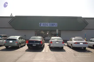 Exterior of a low commercial building with a green sloped awning fascia bearing a white sign reading VEDIC TEMPLE with a purple logo. Four parked cars face the entrance across an open asphalt lot under a pale sky: a silver-green hatchback, a dark sedan with a Cadillac-style rear, a white sedan and a white sedan on the right. Glass storefront doors reveal interior shelves. No people are visible.