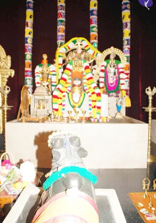 A photograph of a temple-style shrine with a white stone platform holding a central deity murti draped in striped garlands of yellow, red, green and white, with a crown-like headpiece and a tilak on its face. Flanking figures and a smaller shrine structure stand on the platform. Tall pillars wrapped in colourful patterned cloth rise behind. In the foreground a dark stone linga-like form wears a teal garland and a white and gold lei, resting on a low pedestal beside brass oil lamps, flowers, fruit and offering trays. No people are visible.