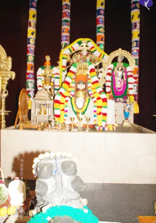Photograph of a decorated altar shrine with a central deity murti draped in multicolored striped garlands and wearing a crown, flanked by two smaller standing figures in similar garlands. Tall pillars wrapped in colorful patterned fabric rise behind the altar. In the foreground, a dark stone object wrapped in a turquoise garland and topped with white flower garlands sits on a counter, with oil lamps, brass lamps, a small shrine canopy, and fruit offerings nearby.