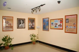 An interior corner of a plain cream-walled room with a beige tiled floor, track lighting and a ceiling dome camera. Several framed posters hang on the walls: at left a poster titled Vedic Rituals and Ceremonies with photo grids, a black-and-white portrait of a smiling woman with long dark hair and a beaded necklace, and a dark framed poster with photo panels. On the right wall, a blue-panelled poster and a multi-panel poster with yellow, blue and pink sections, and a tall poster labelled NITHYA showing a seated figure in meditation and yoga photographs. Two potted plants stand on the floor and a small information stand is at centre. No people are visible.