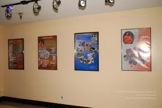 A photograph of a cream-painted wall with four framed poster panels hung in a row beneath track lights. The leftmost panel has an orange background with a grid of small photographs. The second panel is orange with a photo of a figure in orange clothing at top and a cluster of pictures below. The third panel has a blue background with a black-and-white portrait at top and a collage of colour photographs. The rightmost panel is orange and white with a portrait of a figure in orange robes seated at top, a block of text, and a collage of photographs below. No people are visible in the room.