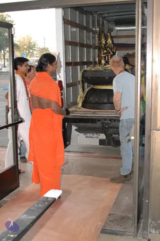 A photograph at a building entrance where a delivery truck's rear doors are open, revealing a wooden-floored cargo interior. A large dark stone pedestal with lotus-petal mouldings sits on a wooden pallet, strapped with yellow lifting straps, with two gilded deity figures standing above it. In the foreground a figure in a flowing saffron-orange wrap, long dark hair, and bare back stands facing the truck. To the left, a man in white clothes and a saffron-edged shawl with beaded mala looks on, and a man in a light grey t-shirt and jeans stands at the open doorway on the right. A white 'Whittier, CA' delivery sign is visible on the truck's side. Two people are visible in addition to the figure in orange.