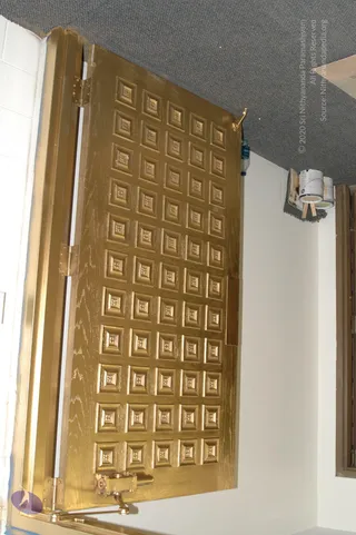 A photograph, shot sideways, of a large gold-painted wooden door with a grid of raised square panels, each carrying a small embossed motif, set in a gold-painted metal frame with a brass latch and hinges. A grey carpeted ceiling and white walls surround it; a bottle of water and two paint cans sit on a shelf near the upper right, and a doorframe edge is visible at the right. No people are visible in the scene.