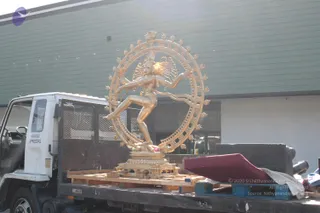 A large gold-coloured metal statue of Nataraja, the dancing Shiva form, stands on a wooden pallet on the flatbed of a white Isuzu FSR truck with DIESEL written on the bed. The dancing deity murti is surrounded by a ring of flames with a raised leg and outstretched arm, set on a tiered pedestal. Behind the truck is a green-and-white corrugated building wall and a glass entrance. Maroon cloth, a dark slab and other packing materials lie on the right side of the bed. No people are visible.