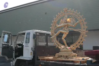 A large gold-coloured bronze statue of Nataraja, the dancing Shiva deity murti, stands on a flatbed truck bed on wooden pallets. The figure is in a dynamic dance pose inside a ring of flames, one leg raised, with multiple arms extended. The white Isuzu FSR truck with an open cab door is parked outside a green corrugated building under bright daylight.