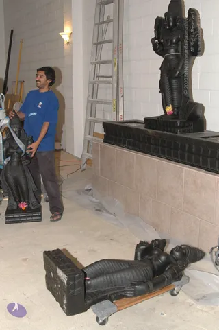 A photograph inside an unfinished building with white cinder-block walls and a light-coloured floor covered in plastic sheeting. A young man in a blue t-shirt and dark trousers stands at left, smiling, with his hands on a black stone deity statue wrapped in a blue strap. Behind him a metal stepladder leans against the wall. At right a tall black stone standing deity murti with multiple arms and a tall crown stands on a black stone pedestal set against a tiled base, with a small yellow and pink flower offering at its feet. In the foreground a large black stone reclining figure lies on a wheeled wooden dolly, its head and feet visible. A small pink flower sits at the base of the left statue.