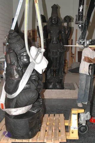 A photograph inside a warehouse-like workspace where dark stone deity murti are being handled. In the foreground a large black stone sculpture of a seated multi-armed deity is suspended by grey and yellow lifting straps, with a worker in a white shirt partly visible behind it. Behind, a standing black stone deity murti with a tall conical crown and multiple arms stands on a pedestal in a recessed base. On the right a worker in a white t-shirt and jeans stands beside a yellow pallet jack and a black crane arm. Wooden pallets and crates lie on the grey floor. No a person candidate is visible.