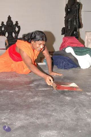 A photograph inside an unfinished temple hall with a grey concrete floor. A man with long dark swept-back hair, wearing an orange shawl over a red garment, leans low on one hand and places a small reddish-gold metal plate on the floor with the other, smiling. Behind him stand dark stone deity murti sculptures, one with multiple arms, and a stack of folded cloths in red, green, blue and white sits against the wall. a person: the long swept-back hair, orange and red saffron-style clothing and visible red tilak mark are consistent with the page's identification, but the appearance is not conclusive. A second partially visible person in white stands at the left edge.