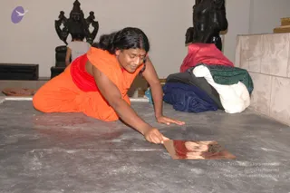 A photograph inside a bare temple-in-progress room with a grey concrete floor and pale tiled wall. A person in an orange saffron garment with a red sash kneels and leans forward, reaching to place a thin copper plate on the floor. Behind them stand dark stone deity murti figures, one with multiple raised arms, and a stack of folded cloths in red, navy, green and white sits against the tiles. A small red tilak mark is visible on the forehead. The scene suggests construction or installation work on a temple space.