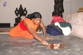 A person in an orange robe and red undergarment kneels on a grey floor in an unfinished temple interior, reaching forward to place a thin copper plate on the ground. The figure has long dark hair pulled back and a small red mark on the forehead. Behind them stand dark stone deity murti figures on pedestals, one with multiple arms, and a pile of folded red, blue, green and white cloths rests against a tiled wall. A small white-clad figure is partly visible in the background. Copper plate placement suggests temple construction work.