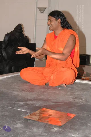 A man with long dark hair and a red bindi sits cross-legged on a grey stone floor, smiling and gesturing with open hands. He wears a sleeveless saffron-orange robe over a red garment. In front of him lies a flat copper plate engraved with a yantra-like geometric pattern. Behind him stands a dark carved stone deity murti with a wall sconce on the pale wall.