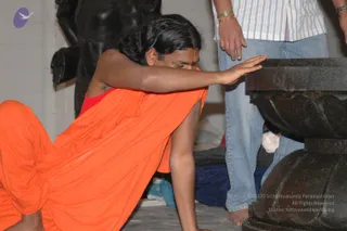A photograph of a man in orange robes kneeling on a stone floor, bending forward with his right hand pressed flat against the top of a dark stone pillar base with a carved lotus-petal profile. He wears an orange wrap over a red undergarment, has a red mark on his forehead and dark hair pulled back. Beside him stands a second person in a white striped shirt, light blue jeans and gold bangles, with a bare foot visible. Behind them a large black stone statue is partly visible against a white wall, and a blue cloth lies on the floor. The scene appears to be an interior temple setting.