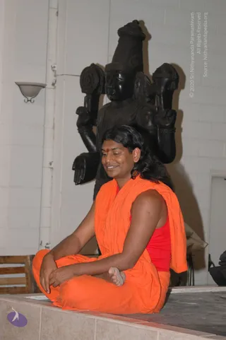 A photograph of a man with long, swept-back black hair sitting cross-legged on a raised tiled platform in a plain white-walled hall. He wears a loose saffron-orange wrap over a red top and has a red bindi on his forehead, his eyes lowered and hands resting in his lap. Behind him stands a large dark wooden deity murti with multiple raised arms and a tall crown. Cues for Swamiji (probable): long swept-back hair, saffron clothing, and a red forehead mark seated before a deity murti, in an in-person setting.