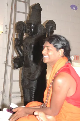 A photograph of a smiling man seated in the foreground, wearing a sleeveless red top, an orange shawl draped over his shoulders and a long rudraksha mala with beads across his chest. His dark hair is swept back, and a red tilak marks his forehead. Behind him stands a large dark stone deity murti with four arms, a tall crown and ornate carved garments, beside an aluminium stepladder against a white block wall. A red EXIT sign is visible at upper right. The seated man's posture and attire are consistent with a visible ceremonial figure, though his identity cannot be confirmed from appearance alone.