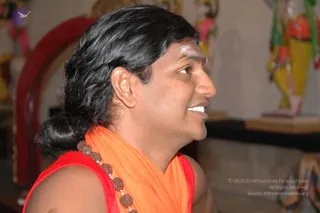 Side profile of a smiling man with long dark hair swept back into a low bun, wearing an orange shawl draped over a red sleeveless top and a strand of rudraksha beads across his chest. A small red tilak mark is visible on his forehead. Blurred deity figures in red and yellow stand in the background near a red archway. Probable Swamiji in person, based on the long swept-back hair, orange shawl and rudraksha mala. Photo taken indoors at a temple setting.