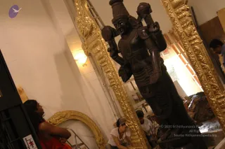Photo inside a workshop or building under construction showing a large dark wooden deity murti standing upright with a tall cylindrical crown and multiple arms holding objects, flanked by ornate gold-painted carved frames. In the foreground at lower left, a woman in a red top sits looking up at the sculpture. Near the base of the statue, a person in a white shirt and cap kneels and another person in white with a cap works beside them. A wall-mounted light and a fire exit sign are visible, with shelving at right.