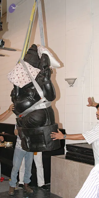 A large black stone deity murti is being lifted by yellow and grey lifting straps in an indoor room with cream cinderblock walls. Padded cloth wraps protect its upper body and head. A man in a white-and-blue striped shirt steadies the base with one hand, and a second man in jeans and a light shirt holds the statue from behind. A third person's hand reaches in from the left edge. A black-draped table holds a brass vessel and a small brass lamp. A tiled platform sits at lower right.