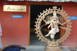 A gilded bronze Nataraja statue dancing within a flaming ring of fire (prabhamandala) stands outside a red brick building entrance. The deity has four arms, a crown with a rearing crest, a striped headcloth, and a garland of pink and white flowers draped over its shoulders and waist. A gold-lettered sign reading "Ananda Sabha" and a blue plate reading "ANANDASABHA" are mounted on the wall, with "NITHYANANDA" partly visible on a second sign to the right. A white cloth and a dark blue cloth lie on the ground at the left edge. No people are visible.