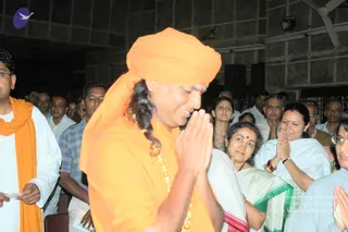 A man in saffron-orange robes and an orange turban stands in the foreground with palms pressed together in namaste, eyes lowered and smiling, a red tilak on his forehead and a gold-toned necklace across his chest. Behind him, a crowd of seated and standing attendees in white and pastel clothing, several with hands folded in greeting, fill an indoor hall with dark textured walls, a ceiling fan and speakers. A man in a white kurta with an orange shawl stands at left holding a small paper. A woman in a white sari with a green and red border and a red bindi stands at center. The setting appears to be a satsang or public gathering.