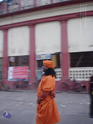 A figure in a saffron-orange turban and long orange robe walks in profile along a paved street past a red-and-cream colonnaded building. The figure has long dark hair falling below the turban, and the orange robe is draped over one shoulder with a rust-coloured garment visible beneath. Hands are held together in front. Behind them, a red-framed signboard and two parked bicycles line the street, and a small red-and-white sign is mounted on the building. The image is motion-blurred. Swamiji (probable): long swept hair, saffron turban and orange robes are visible cues, shown walking as a sannyasi in a public street.