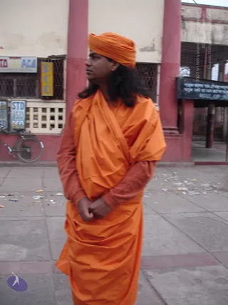 A man in a saffron-orange turban and matching orange wrap-around robe stands on a paved street in front of a pink-columned building with shuttered shopfronts. He has long dark hair falling past his shoulders, a darker rust-coloured inner garment visible at the arms, and his hands are clasped in front of him as he looks off to one side. Behind him a bicycle leans against the wall beside signage for STD/ISD/PCO telephone booths, and scattered litter lies on the ground.