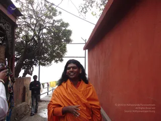 A photograph of a long-haired man in an orange-saffron shawl walking along a narrow path beside a tall red-painted wall, smiling with a red bindi on his forehead. He is wrapped in a saffron cloth over his chest and keeps one hand near his stomach. Behind him a man in a dark jacket and jeans stands near a tree with pale branches, and washed yellow cloth hangs on a railing. A partially visible hand in white clothing and a camera strap appear at the left edge.