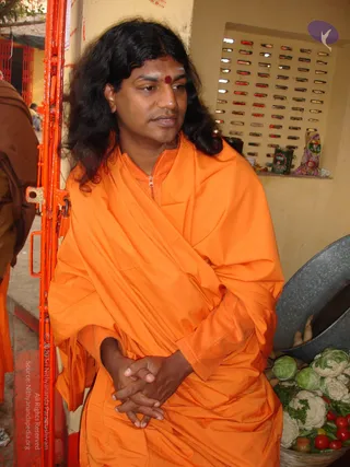 A photograph of a long-haired man in a saffron-orange sannyasi wrap and zipped orange top stands beside an orange metal gate, hands clasped in front of him. He has a red bindi on his forehead and sits in front of a wall with a grid of small mirrors. In the foreground at right, a metal basin holds fresh cauliflower, cabbage, leafy greens and red tomatoes. A person in brown robes is partly visible at the left edge. Swamiji (probable): long swept-back hair, saffron-orange ceremonial wrap and a red forehead mark.