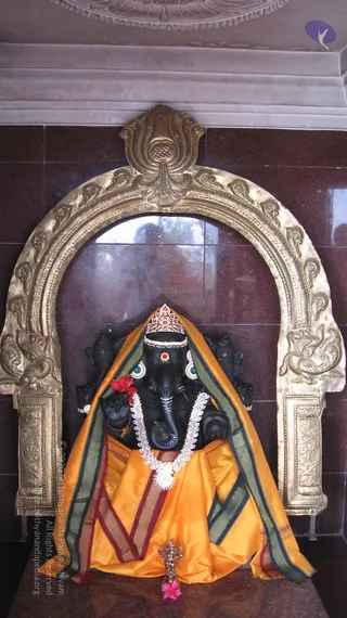 A black stone Ganesha murti sits within an ornate brass arch set against a dark maroon granite wall. The deity wears a silver and red jewelled crown, a white beaded garland, and an orange-and-green draped cloth, with a small brass figure and a pink flower placed at its base. Red flowers rest near its shoulder, and a carved ceiling motif appears above the arch.