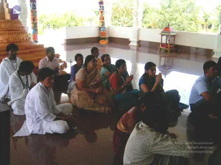 A photograph inside a pillared open hall with a polished reflective floor, where a group of devotees sits on the ground with hands pressed together in prayer or meditation. A woman in a beige sari with a gold border sits at centre, next to a woman in a red and green sari and a woman in a dark blue top with folded hands. A man in a white shirt sits at left near a golden pedestal, and a young man in white with a mala leans at the far left. Behind them are a small red-and-gold shrine and green trees through the open arches. Several more seated people in white and pastel clothes are visible at the right and foreground.