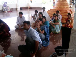 Indoor photo of a group of devotees seated on a polished reddish-brown marble floor in a temple hall, with a tall orange-gold stepped pedestal behind them. In the foreground a man in a light blue shirt and dark trousers sits cross-legged, and a woman in a blue sari sits with palms pressed together in prayer. Beside her a woman in a teal sari and a man in white kurta sit close by, and a small child in red sits at the right edge. Other seated devotees, one in a red sari at far left, sit around a floor drawing of white kolam lines. Framed portraits and a small silver vessel rest on a shelf at upper left, and a candle stand is visible at the right. Total physical people visible: about 11.