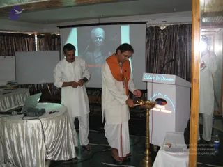 Indoor meditation programme hall with a projector screen showing a black-and-white image of a man in a white turban and dark clothing, partly obscured by the projected words of a title. A man in white kurta and white dhoti with a saffron-orange shawl and a lanyard lights a brass oil lamp at a wooden podium labelled Cidade De Daman and Hotel Lodge text. Beside him stands another man in a white kurta and white trousers, looking down at a laptop on a draped table with a projector and microphone. Bottles of water and books are visible, and a large mirror reflects the room on the right.