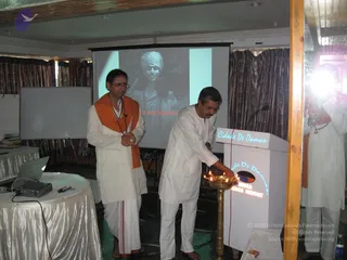 Indoor event photo in a conference hall with dark curtains and a white projector screen. A man in white kurta and white dhoti lights a brass oil lamp (deepam) on a lectern labelled "Cidade De Daman" and "Bhakti Beach Resort", with several small flames burning. Beside him stands a man in a white kurta and dhoti with an orange-saffron shawl draped over his shoulders and a rudraksha-style mala, with his hands folded. The projector screen behind shows a black-and-white image of a smiling person in a turban with the partial caption "...t And Inspira...". A laptop and projector sit on a draped table to the left, and a cameraperson is partly visible at the right edge.