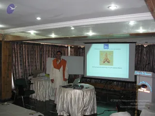 A man in white ritual clothes with an orange saffron shawl draped over his shoulders stands smiling beside a small table with a laptop and projector in a wood-panelled meeting room. Behind him a projection screen shows a slide reading "I Welcome you with all my love & respects" with praying hands and the line "And call upon the blessings of my Enlightened Master". A whiteboard, draped curtains, a brass oil lamp and a lectern with a sign are visible, and a partially visible person sits at the lower right. The scene resembles an introductory talk or meditation programme session.