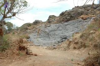 A photograph of a dry, rocky hillside with a bare grey rock face sloping up on the right and a dusty dirt track running up the centre foreground. Scattered loose stones, reddish earth, dry grass and shrubs cover the slopes, and a tree with bare branches stands at the left edge under a pale sky. No people or ritual activity are visible; the scene is an empty rural landscape.
