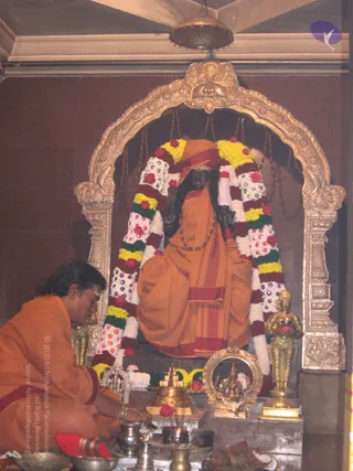 A photograph inside a shrine niche with an ornate golden arch topped by a lion-face motif. A dark stone or metal murti of a seated sage figure in orange-saffron robes, with a saffron turban, rudraksha mala, and long hair, is decorated with thick garlands of white, red, yellow and green flowers. A man in orange robes sits cross-legged at lower left, bent over offerings, apparently performing a puja. Brass and silver ritual vessels, a brass kalasha, a small golden deity figure and a flame-lit lamp sit on the altar in front of the murti. The scene is a puja at the shrine.