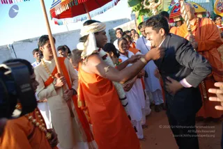 Outdoor procession scene on a dusty ground beside a concrete wall. In the foreground a young man in an orange-saffron robe with a white turban and a white flower garland shakes hands with a smiling man in a dark suit and white shirt. A young man in a cream sherwani with an orange sash holds a long wooden pole of a ceremonial umbrella, and a red and yellow canopy stands above. Behind them people in white kurtas and orange robes line the path, and a man in orange and a man in white stand to the right. A camera operator in an orange robe is at the left edge.