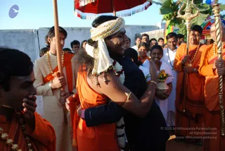 Outdoor procession scene under bright sky. A central figure with long dark hair wears an orange sleeveless garment, a white and gold turban-style headdress with tassels, and a white flower garland. He is embraced from the front by a person in a dark navy shirt with a white garland. Behind them a red and white parasol with fringe is held up, and a woman in white holds a brass pot with a green leaf offering. Two men on the right wear orange robes and gold-toned necklaces, one holding a tall golden trident-topped staff. A man on the left in cream sherwani with orange sash holds a wooden pole with a red umbrella. Crowd of people stands behind a concrete wall.